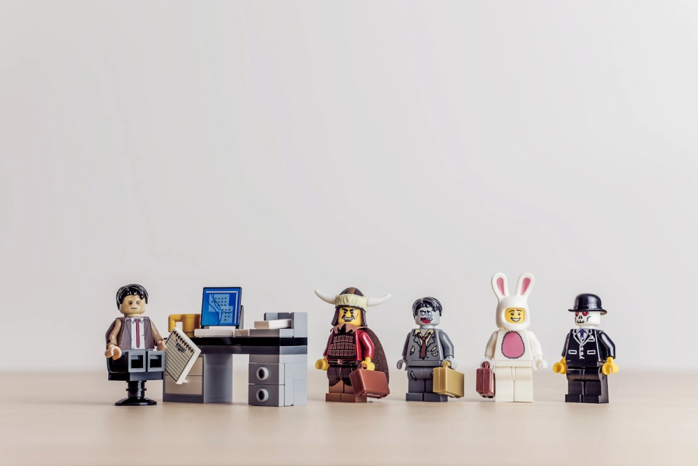 LEGO minifigures in a wide variety of costumes waiting in line in front of an office desk.